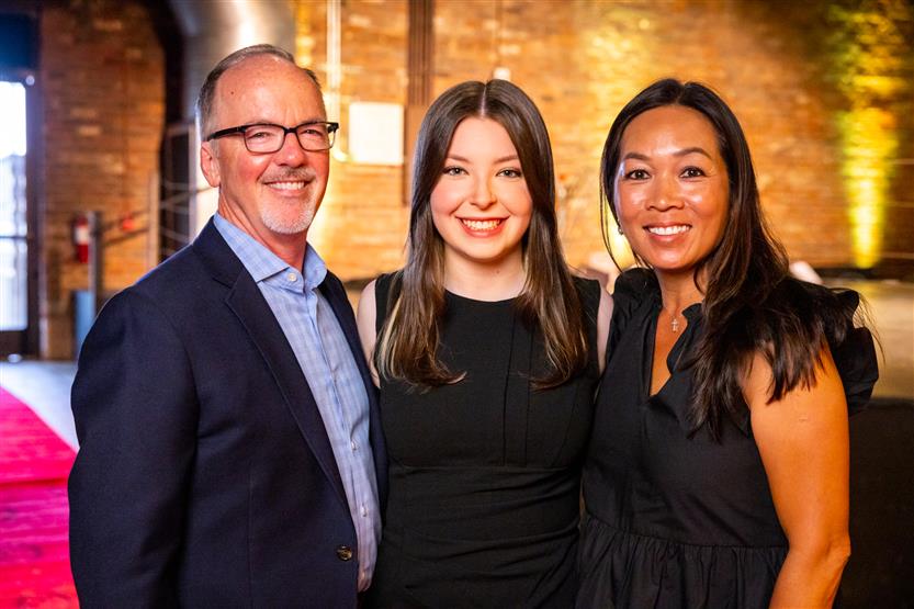 Two women wearing black dresses smile with a man wearing a navy blazer
